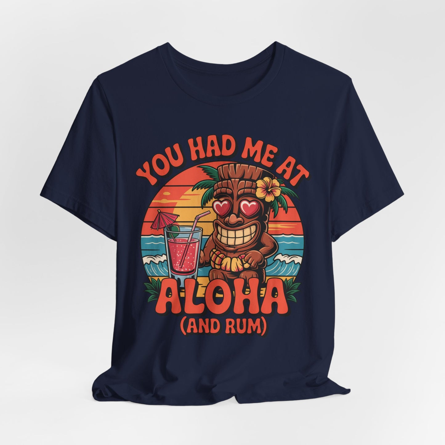 The Tropical Aloha Graphic Tiki T-Shirt for Beach Lovers in navy blue shows a smiling tiki with heart eyes holding a pink drink before a sunset beach, with “You had me at Aloha (and rum)” text—perfect island vibes!.