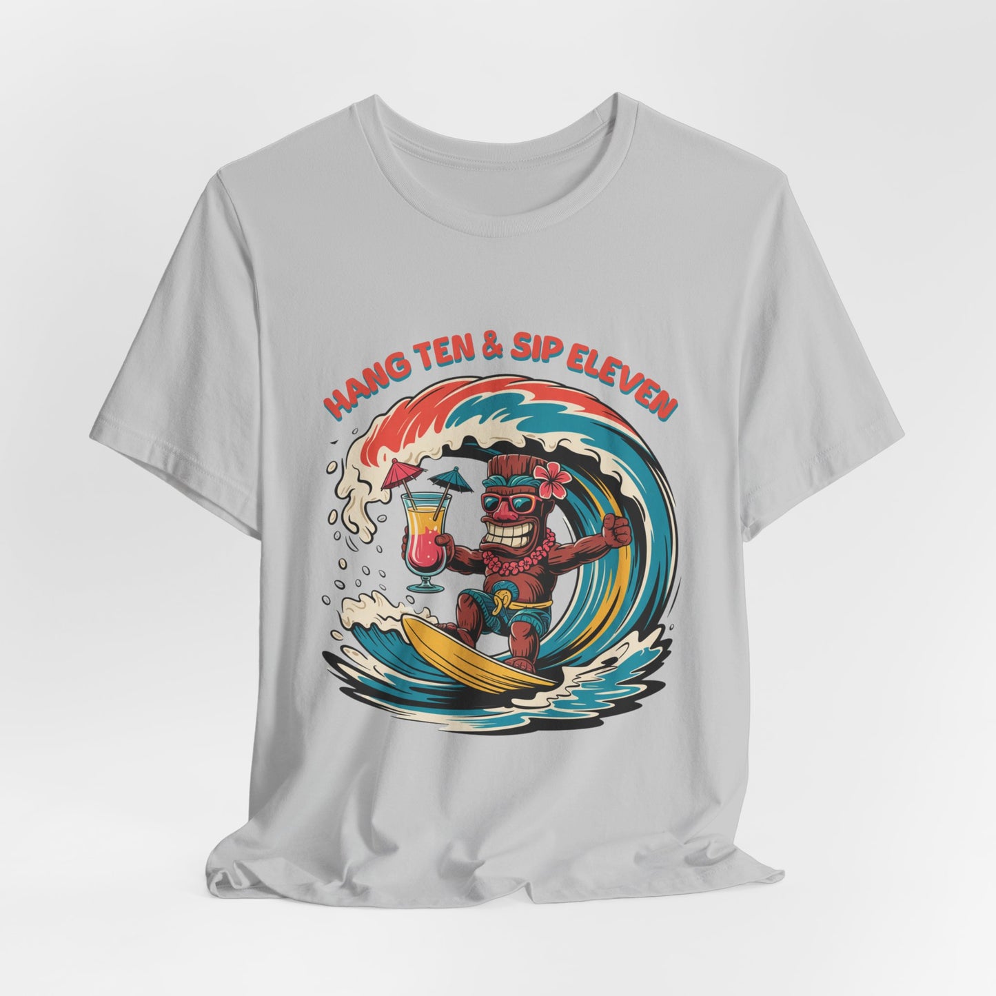 The Surf Vibe Tiki T-Shirt - Hang Ten & Sip Eleven Unisex Jersey Short Sleeve features breathable cotton and a surf-inspired cartoon tiki surfing with a tropical drink, plus orange curved text: HANG TEN & SIP ELEVEN.