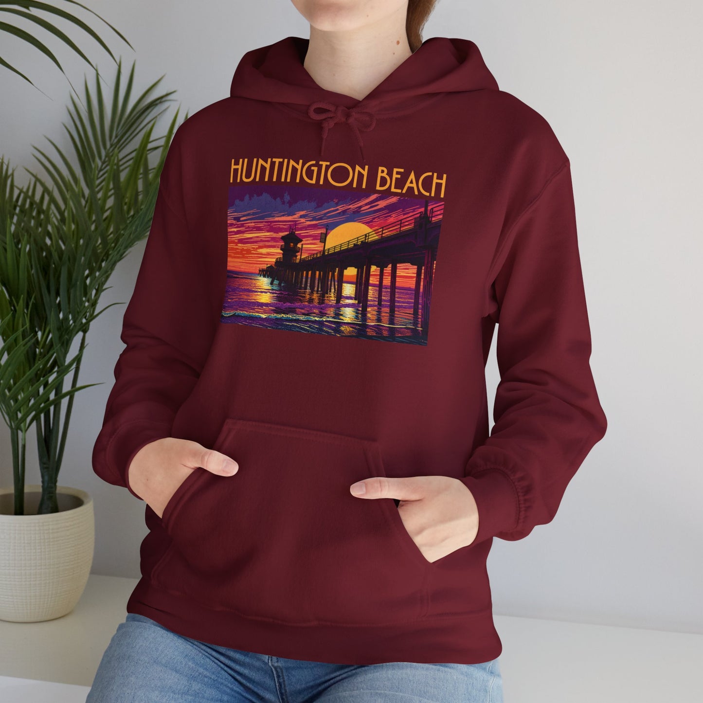 Someone stands indoors by a green potted plant wearing the Huntington Beach Sunset Hoodie | Unisex Heavy Blend™ Sweatshirt, featuring a cozy maroon design with a vibrant pier-at-sunset graphic and Huntington Beach text—ideal casual wear.