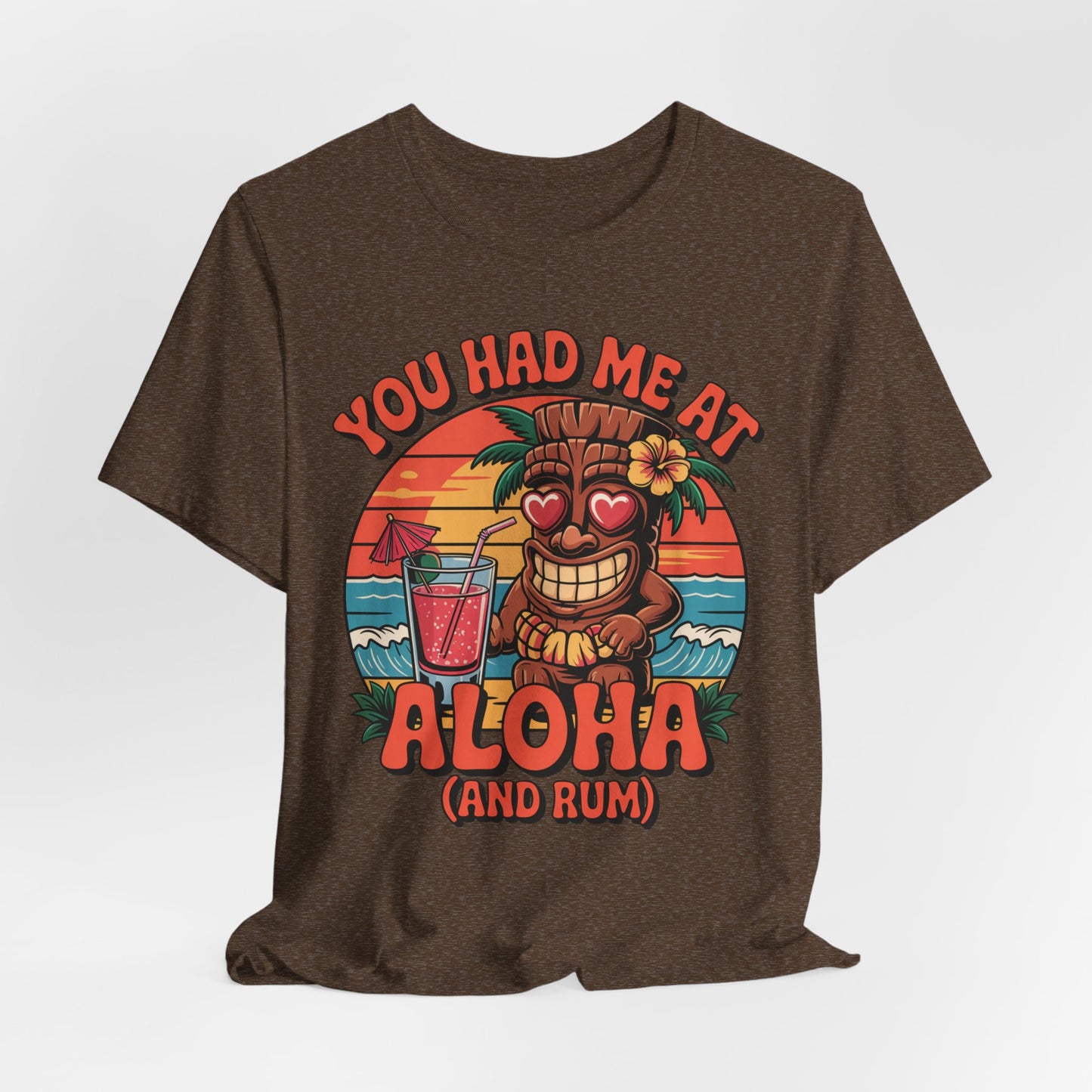 The Tropical Aloha Graphic Tiki T-Shirt for Beach Lovers shows a cartoon tiki with heart eyes holding a drink at sunset, plus You had me at Aloha (and rum)—perfect island vibes for your next beach day!.
