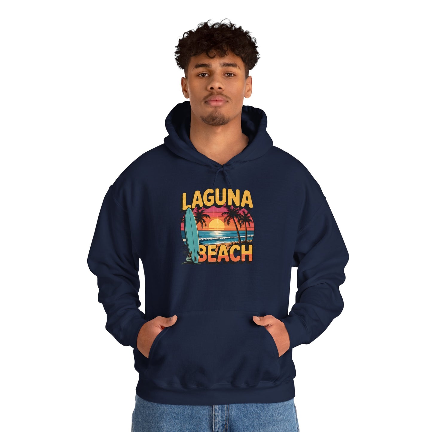 A young man wears the Laguna Beach Sunset Hoodie for Beach Lovers—a navy blue unisex sweatshirt featuring retro palm trees, a sunset, and surfboard design—with hands in the front pocket against a plain white background.