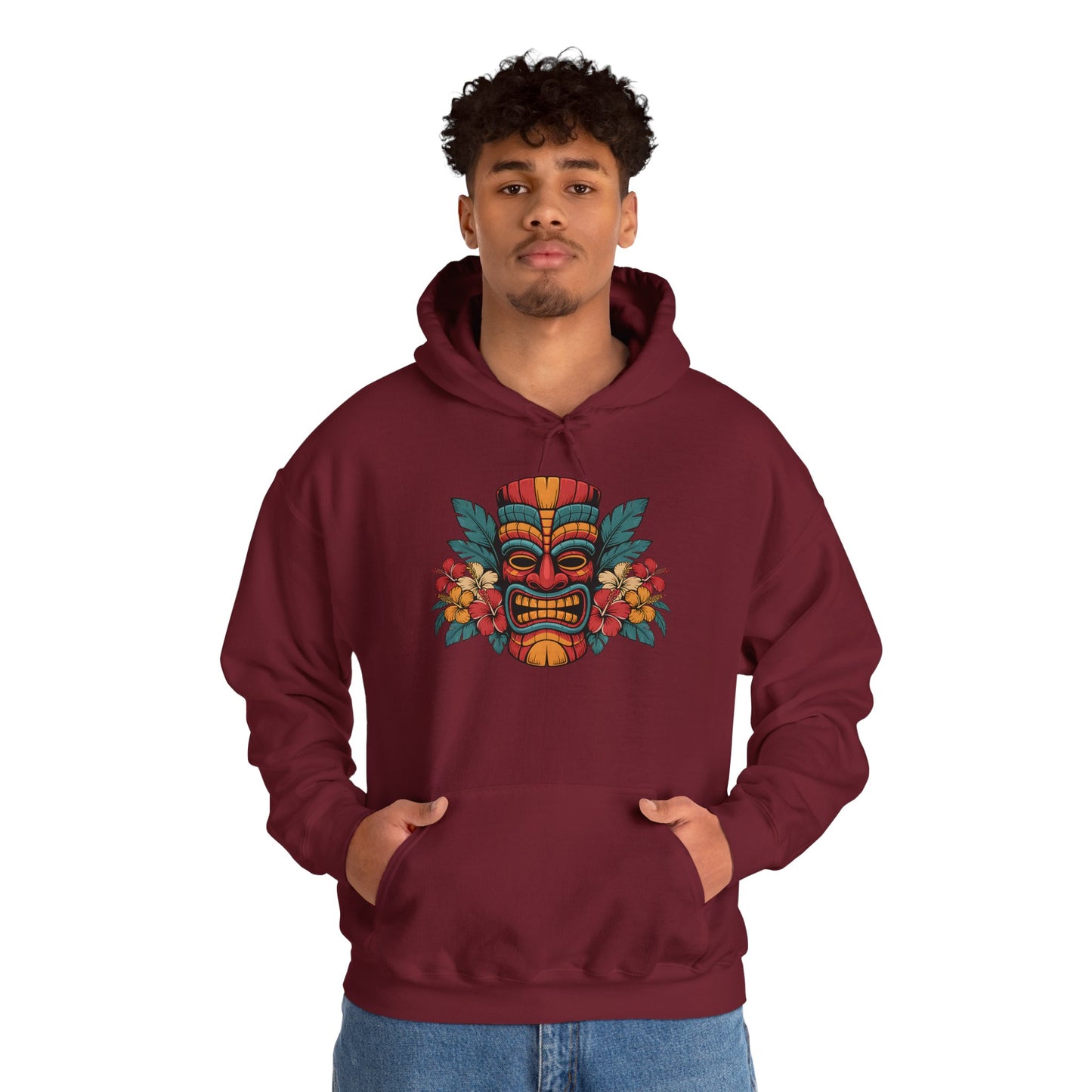 A young man stands facing forward with hands in pockets, wearing the Tropical Tiki Hooded Sweatshirt - Unisex Heavy Blend™, which features a vibrant tropical-inspired design with a colorful Tiki mask and florals, against a plain white background.