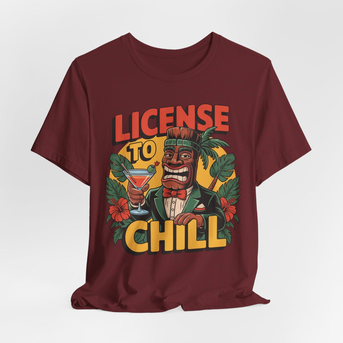 The T-Shirt - License to Chill Tropical Graphic Tiki T-Shirt is a maroon unisex tee with a cartoon tiki in a suit holding a cocktail, tropical leaves and flowers, and LICENSE TO CHILL text—an ideal playful gift for summer barbecues.