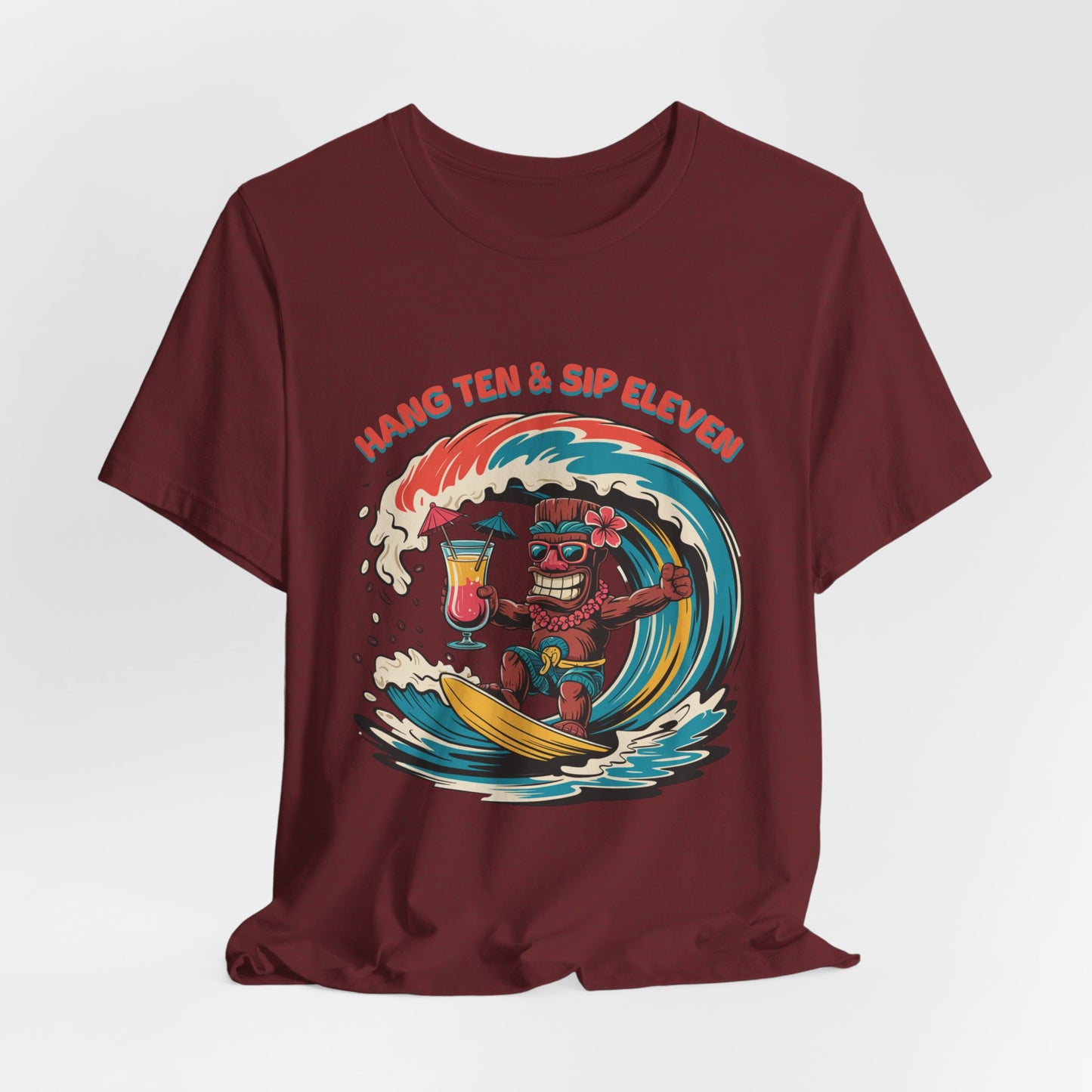 The Surf Vibe Tiki T-Shirt - Hang Ten & Sip Eleven Unisex Jersey features a maroon color with a cartoon tiki surfing a wave, holding a tropical drink. Made of breathable cotton, it has HANG TEN & SIP ELEVEN in bold curved text above.