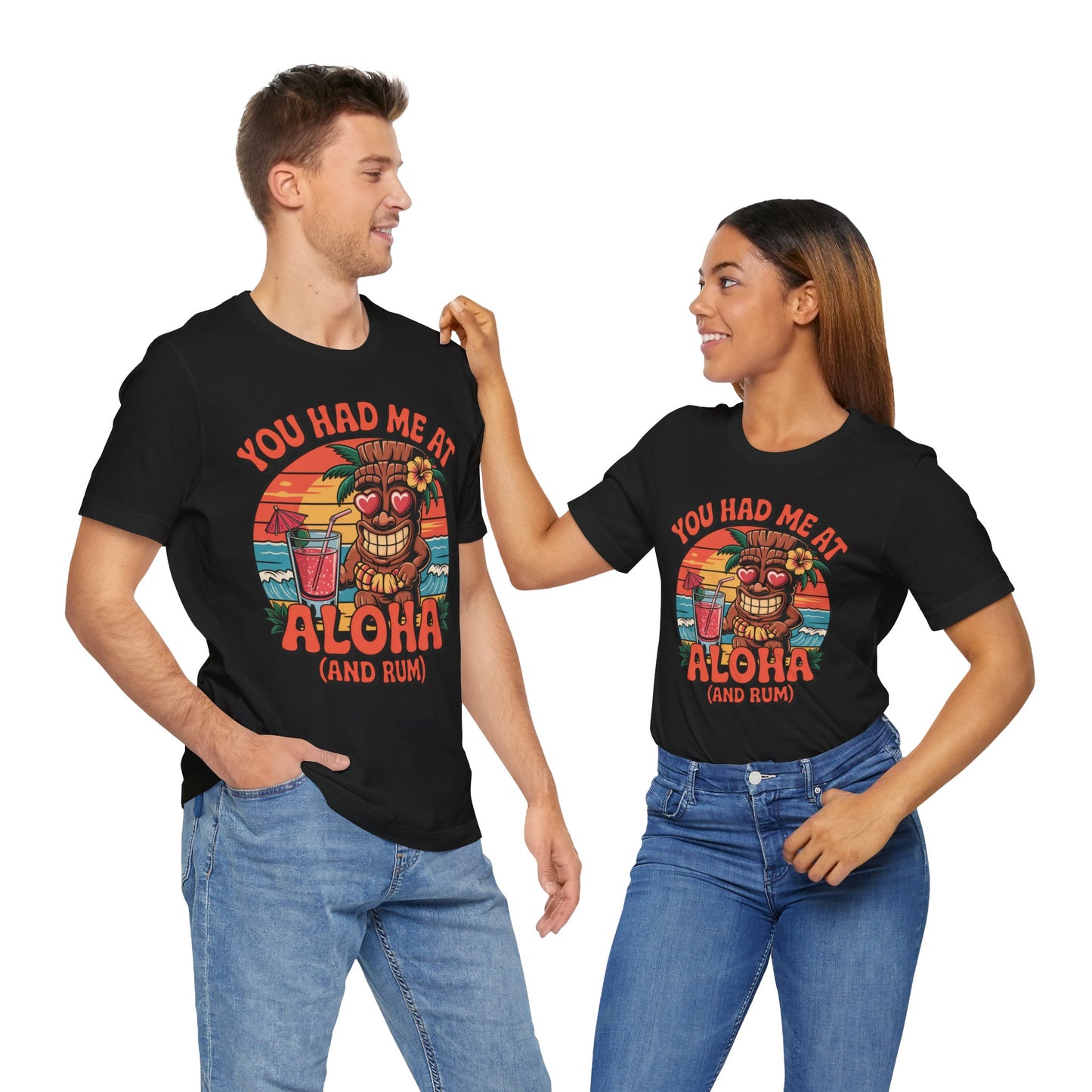 A man and woman smile at each other, both wearing matching Tropical Aloha Graphic Tiki T-Shirts for Beach Lovers with a tropical sunset and You had me at Aloha (and rum)—the woman playfully points at the man, capturing carefree island vibes.