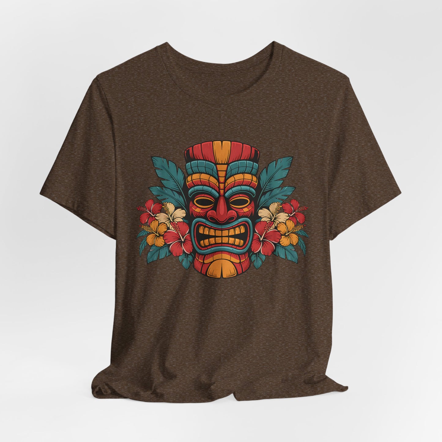 The Tropical Tiki Mask T-Shirt - Unisex Jersey Short Sleeve features a brown tee with a vibrant tiki mask design accented in red, yellow, blue, and surrounded by tropical flowers and green leaves for an island vibe.