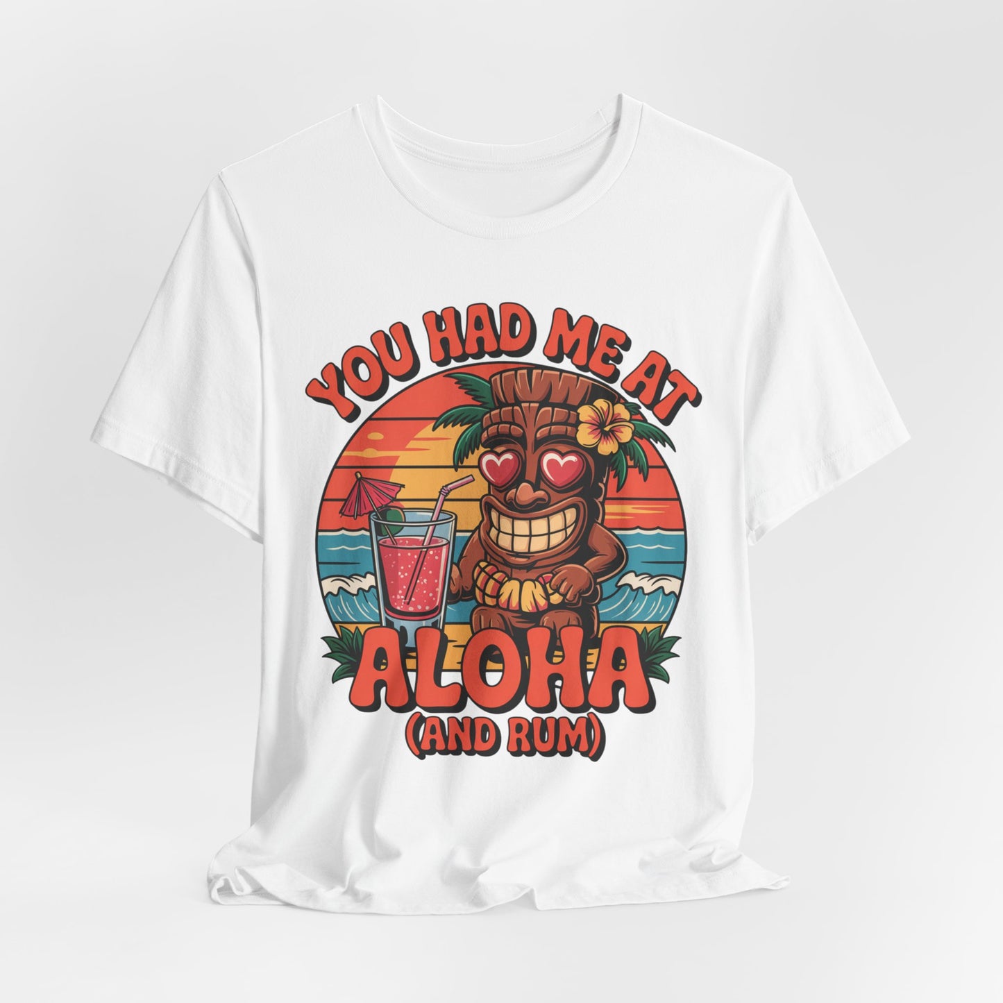 The Tropical Aloha Graphic Tiki T-Shirt for Beach Lovers features a smiling tiki with heart eyes, palm trees, a sunset, and the phrase YOU HAD ME AT ALOHA (AND RUM)—ideal for anyone who loves island vibes.