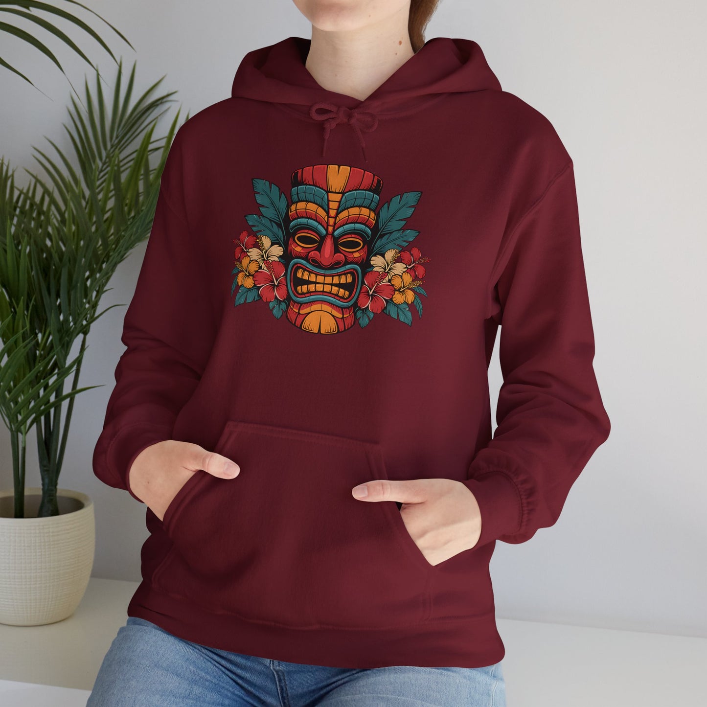A person in a maroon Tropical Tiki Hooded Sweatshirt – Unisex Heavy Blend™, featuring a vibrant tribal mask and flower with a tropical theme, stands indoors near a potted plant—ideal for cozy evenings.