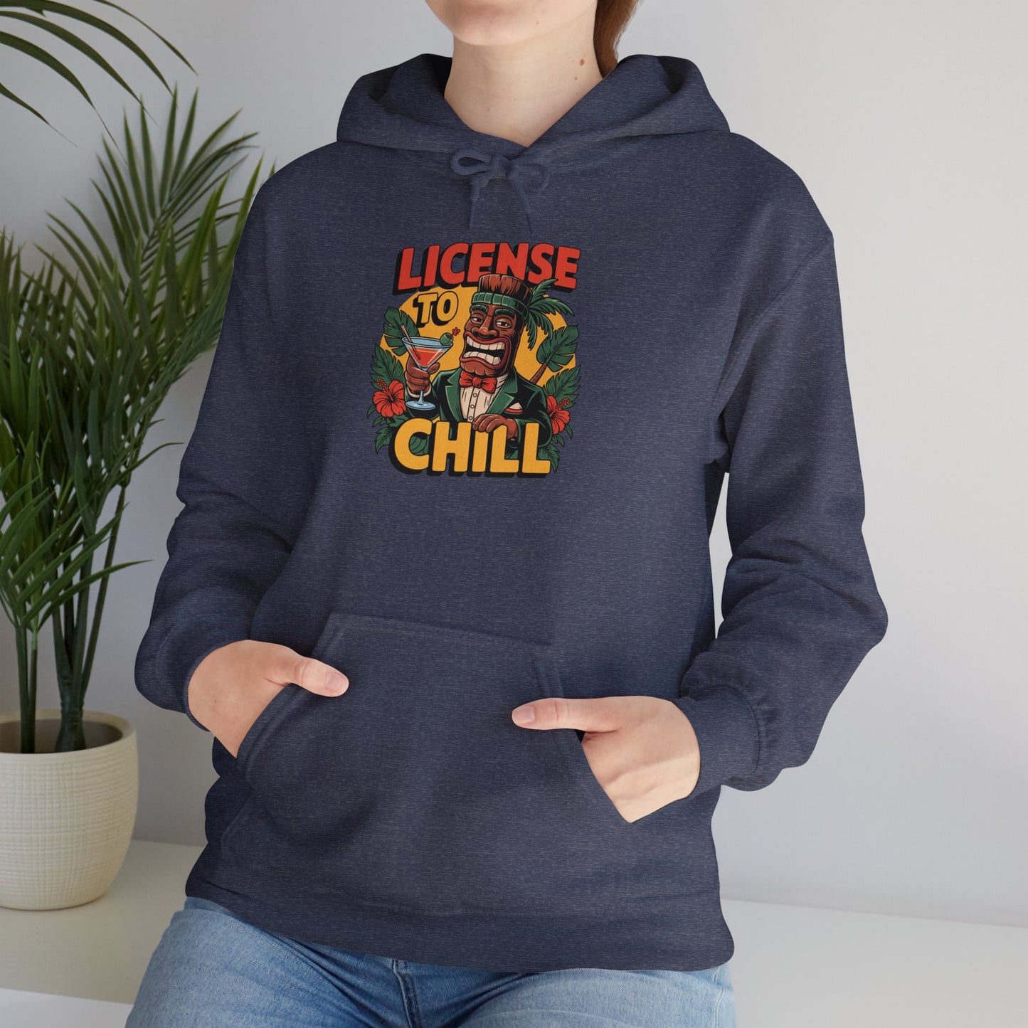 Someone wearing the License to Chill Hoodie - Unisex Heavy Blend Sweatshirt for relaxation and fun, featuring a vibrant tiki face, tropical leaves, and License to Chill text, stands indoors by a green potted plant—a versatile wardrobe essential.