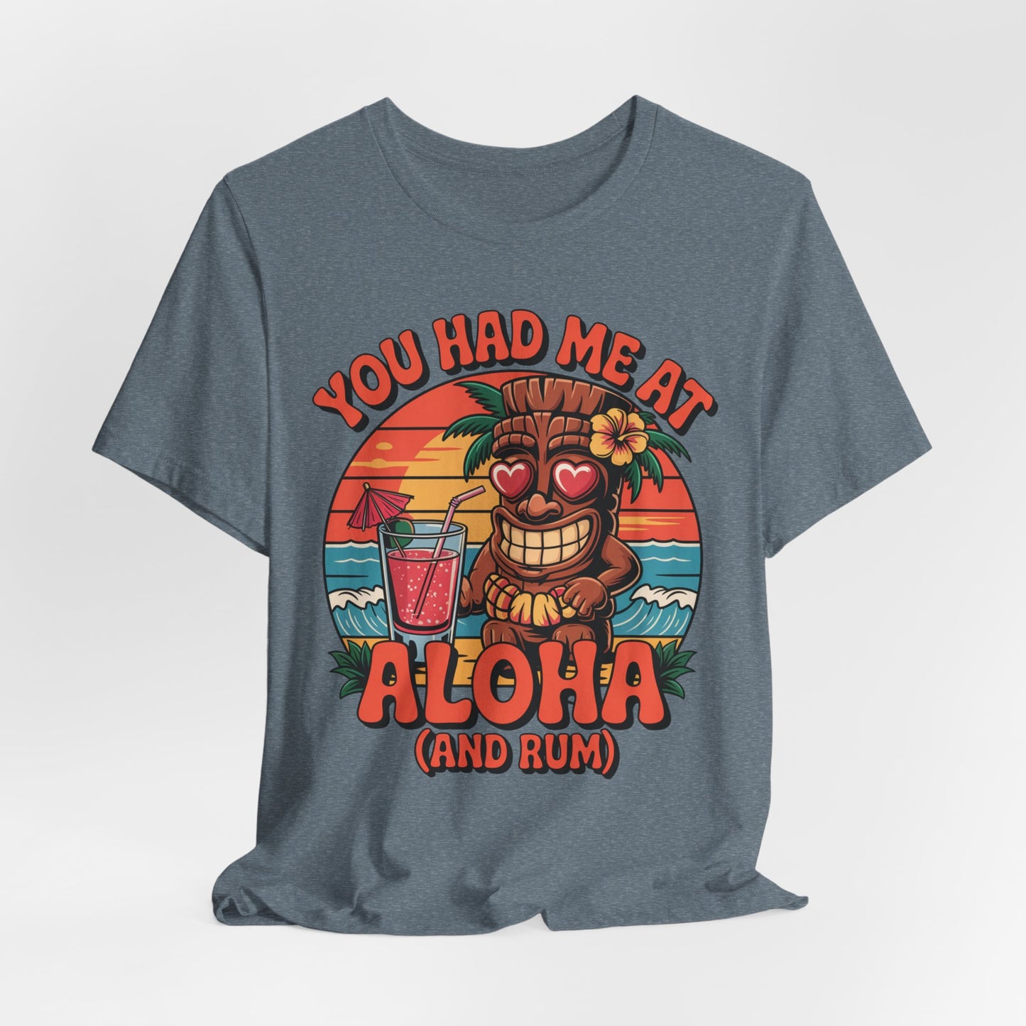 The Tropical Aloha Graphic Tiki T-Shirt for Beach Lovers features a smiling tiki statue with heart-shaped eyes, a cocktail, and a sunset beach scene. The shirt reads: You had me at Aloha (and Rum). Perfect for radiating island vibes!.