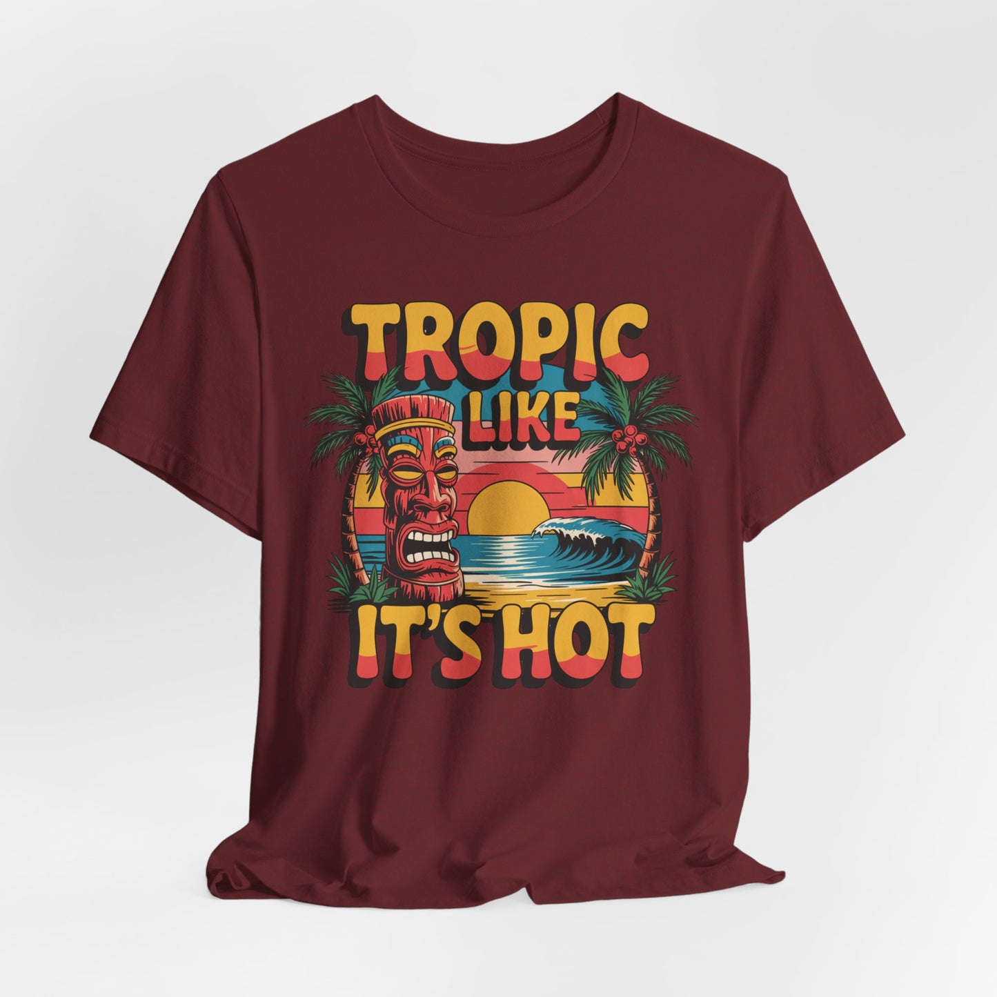 The Tropic Like Its Hot Unisex Tiki T-Shirt features a vibrant tiki face, palm trees, ocean waves, and sunset, with bold yellow and orange “TROPIC LIKE ITS HOT” text—your perfect shirt for summer festivals and vacation vibes.