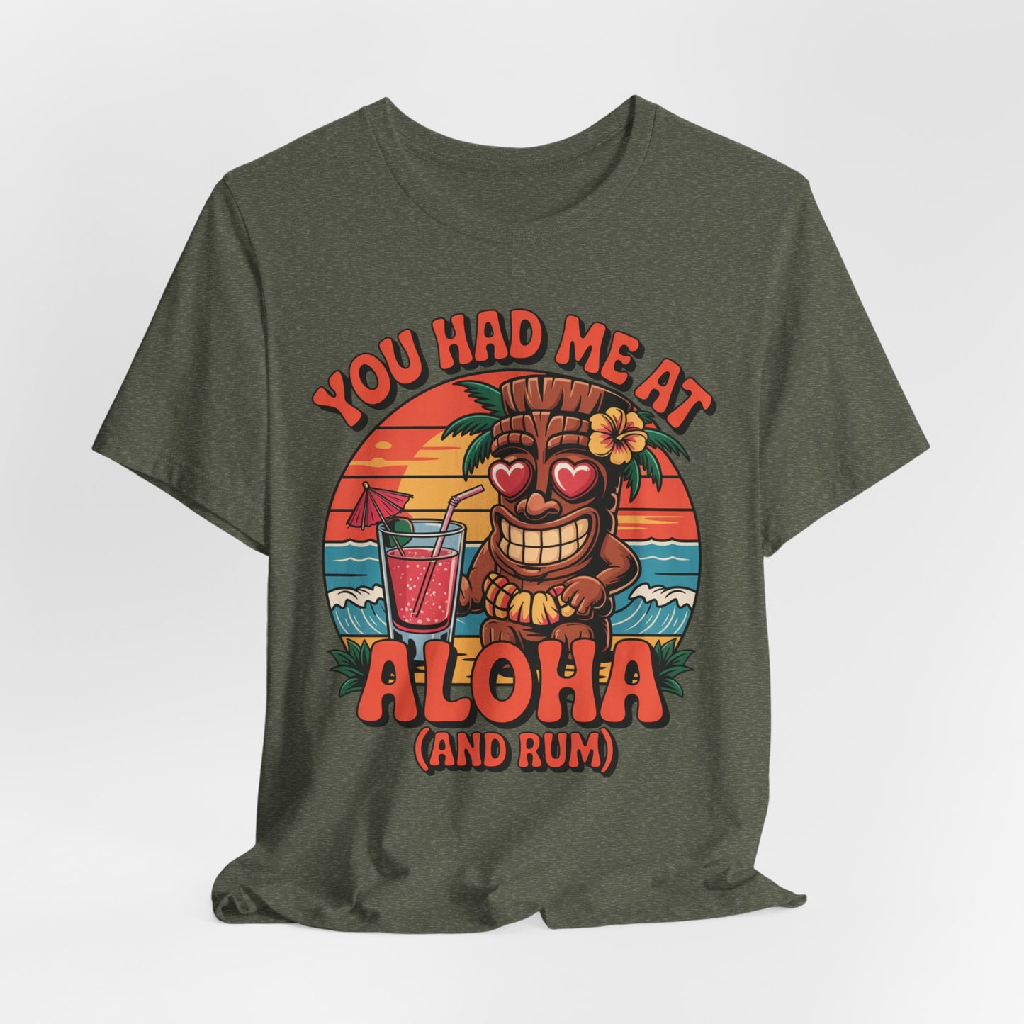 The Tropical Aloha Graphic Tiki T-Shirt for Beach Lovers features a smiling tiki with heart eyes, a cocktail, waves, and a sunset. Bold colorful text reads: You had me at Aloha (and rum), perfect for island vibes.