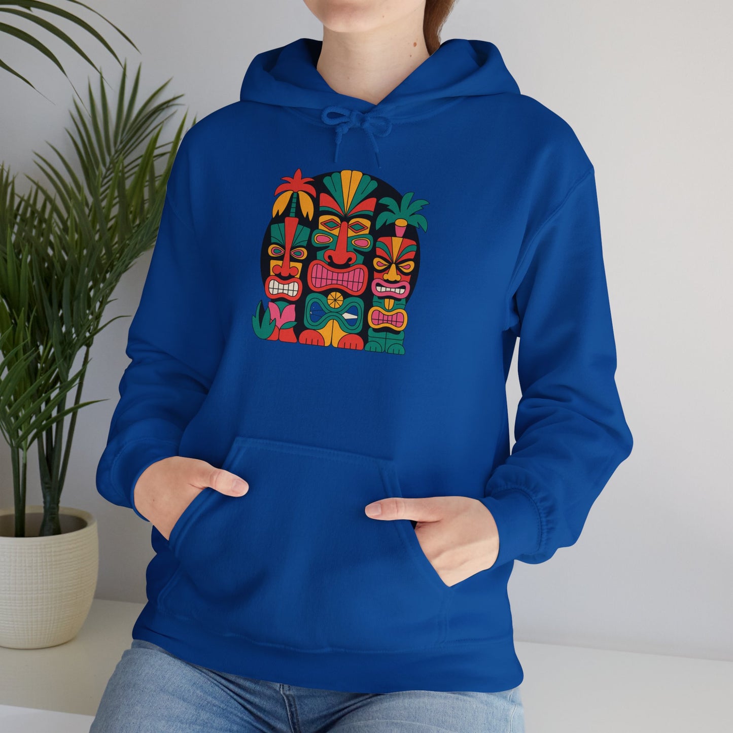 A person in the Tropical Vibe Tiki Hooded Sweatshirt - Unisex Heavy Blend™, featuring a colorful abstract tribal face and cozy kangaroo pocket, stands by a white table and potted green plant.