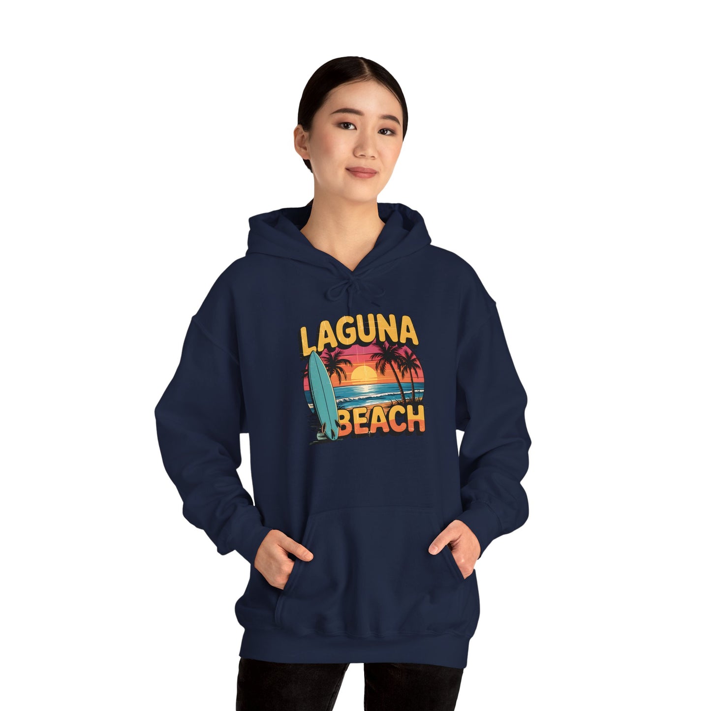 A person smiles with hands in the front pocket, wearing the Laguna Beach Sunset Hoodie for Beach Lovers in navy blue, featuring a colorful sunset, palm trees, surfboard, and Laguna Beach text against a plain white background.