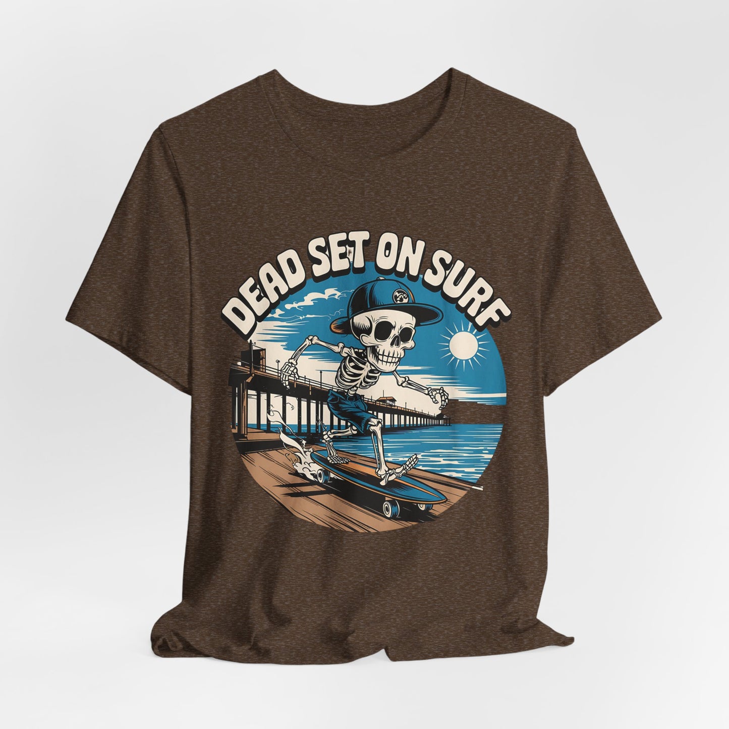 The Skateboard Skeleton Unisex T-Shirt - Dead Set on Surf Design features a cartoon skeleton in a cap skateboarding by a beach pier, with waves, blue sky, sun, and “DEAD SET ON SURF” text above. Brown color.