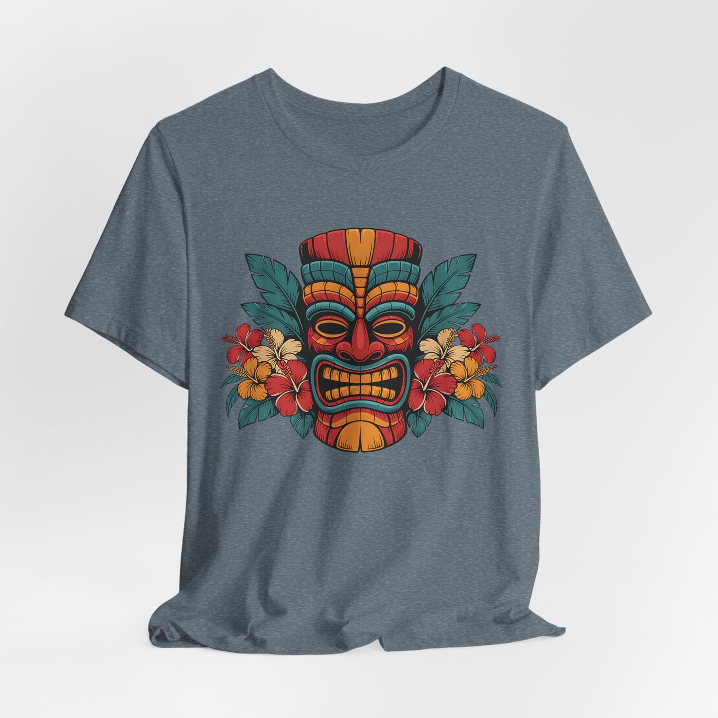 The Tropical Tiki Mask T-Shirt - Unisex Jersey Short Sleeve features a vibrant tiki mask graphic surrounded by red, yellow, and orange tropical flowers with green leaves on a grey shirt, laid flat on a light background.