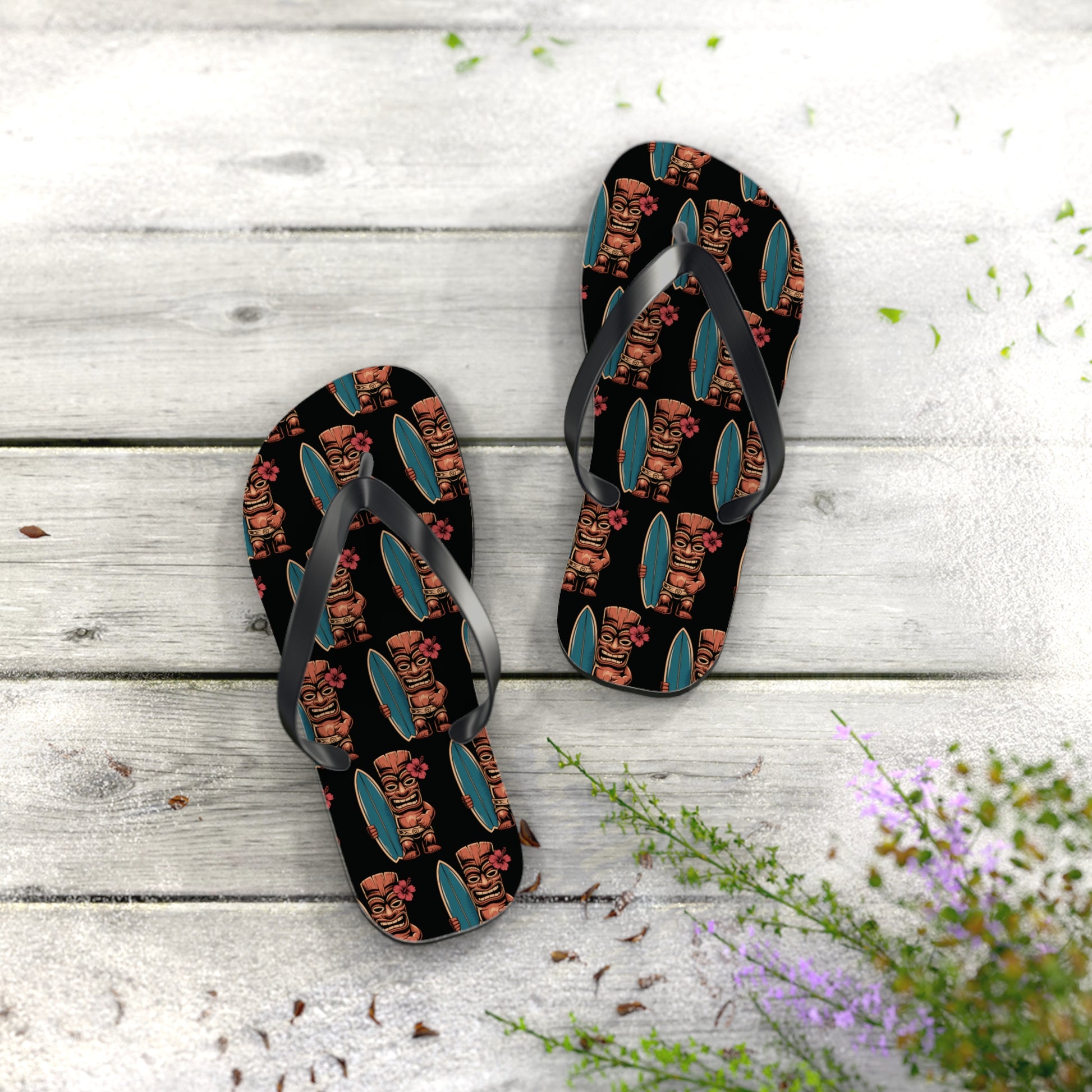 The Tiki Surf Flip Flops - Fun Summer Beach Sandals, with vibrant tiki faces and teal surfboard prints, rest on a rustic wooden surface accented by green foliage and small purple flowers in the corner.