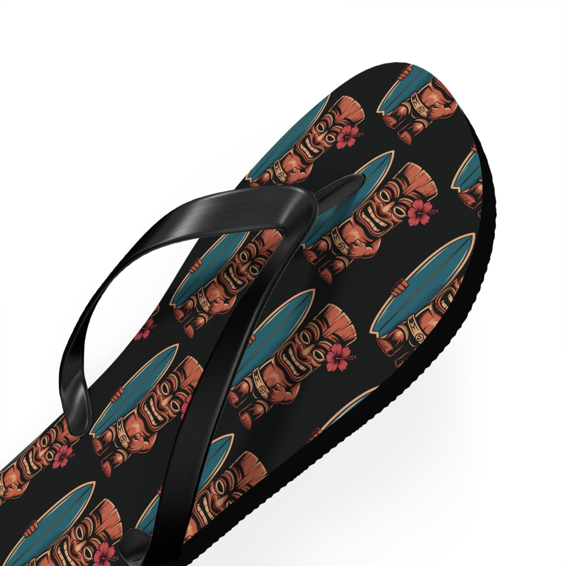 The Tiki Surf Flip Flops - Fun Summer Beach Sandals feature a black base with lively brown tiki faces, pink flowers, and blue surfboards, plus black straps and a soft polyester suede finish. Displayed on a white background.