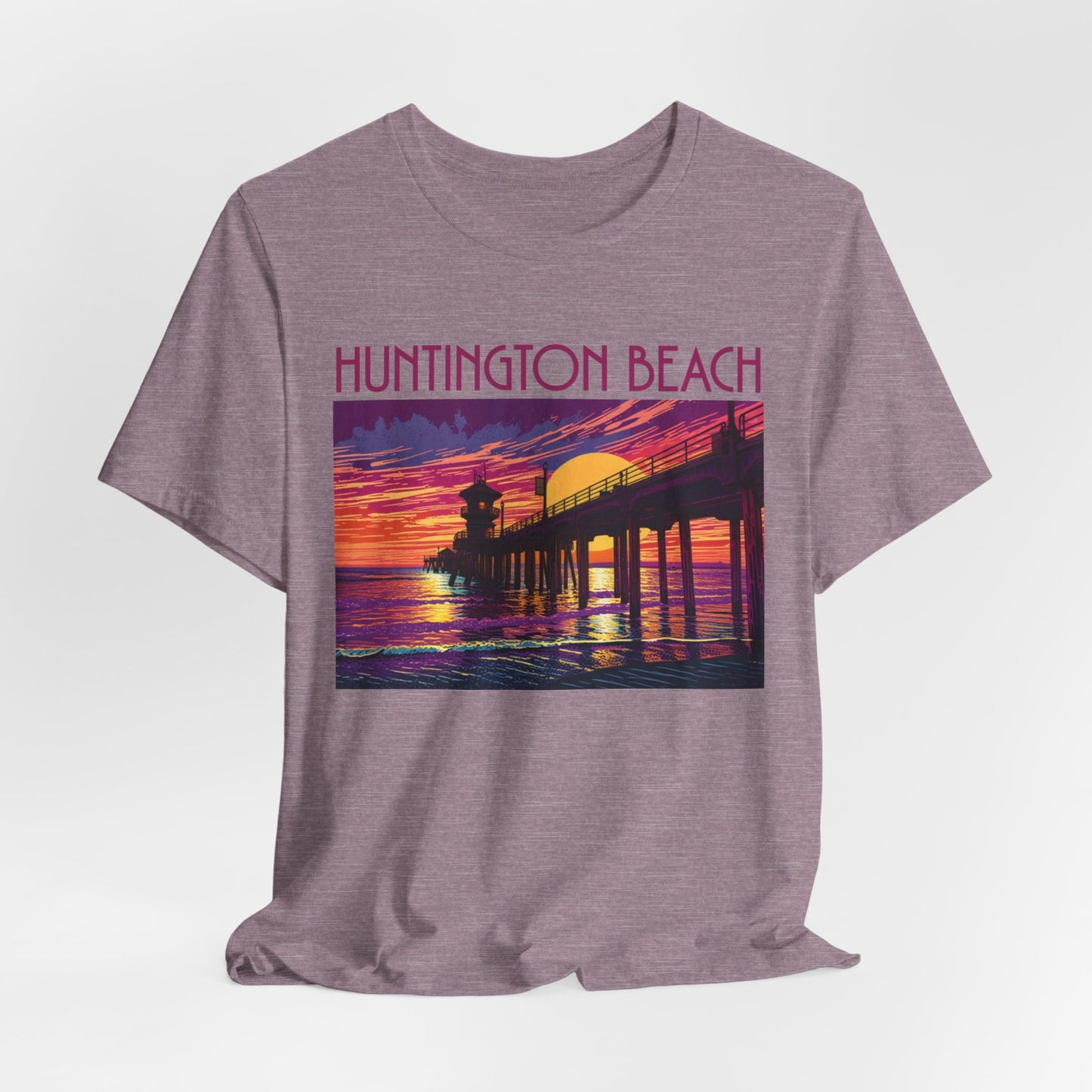 The Huntington Beach Sunset T-Shirt – Unisex Jersey Short Sleeve features a vibrant pier-at-sunset graphic with orange, purple, and pink tones, plus bold pink “HUNTINGTON BEACH” text to capture the local beach culture.