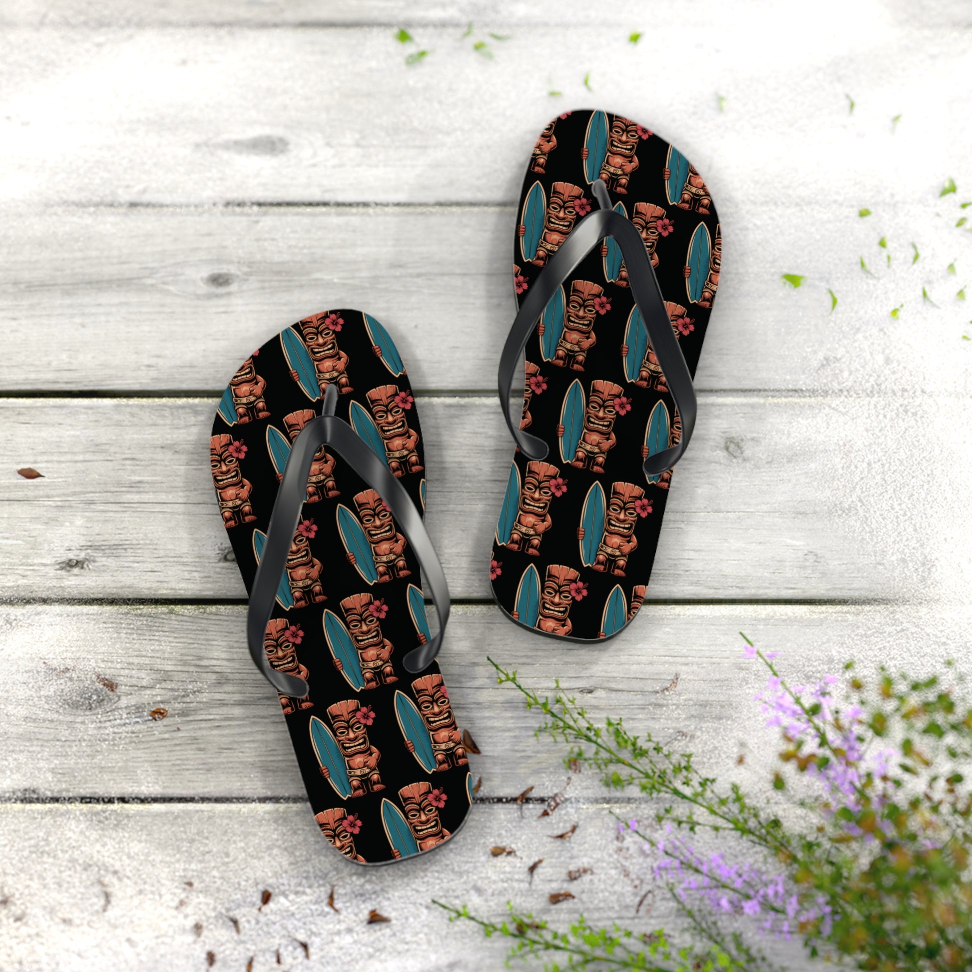The Tiki Surf Flip Flops - Fun Summer Beach Sandals feature a black base with colorful cartoon tiki faces and surfboards, set on a weathered white wooden surface accented by green plants and purple flowers in the corner.