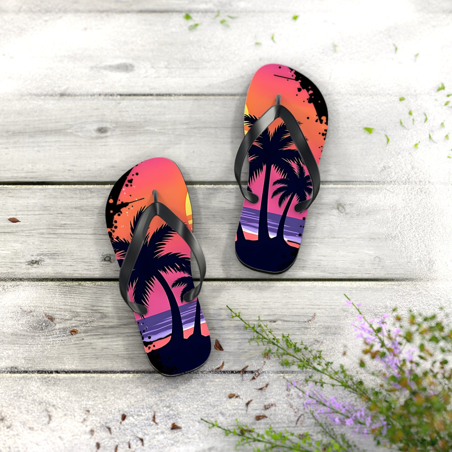 The Tropical Sunset Flip Flops - Beach Vibes Sandals for Summer Fun, featuring a sunset and palm tree design, sit on a light wooden surface with green plants and purple flowers nearby—ideal for your next beach trip.