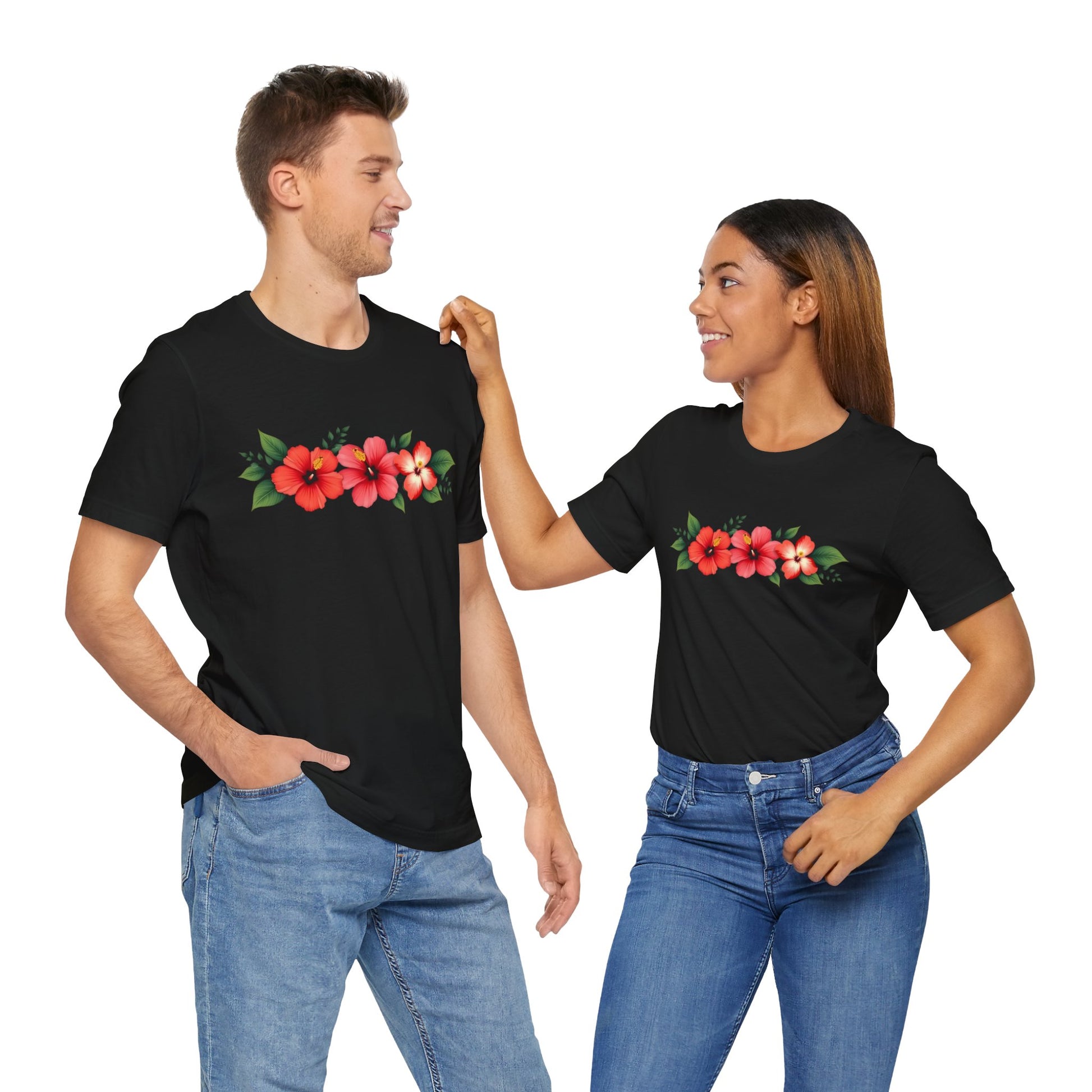 A man and woman smile at each other in matching black Floral Unisex Short Sleeve T-Shirts—perfect for spring and summer—with a red hibiscus design, paired with blue jeans, as she touches his shoulder.