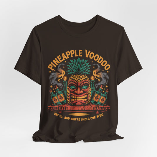 The Pineapple Voodoo Tiki T-Shirt is a black unisex jersey featuring a vibrant tiki mask with pineapple top, surrounded by bottles and smoke. “Pineapple Voodoo” is above, “One sip and you’re under our spell” below. Perfect for beach lovers!.