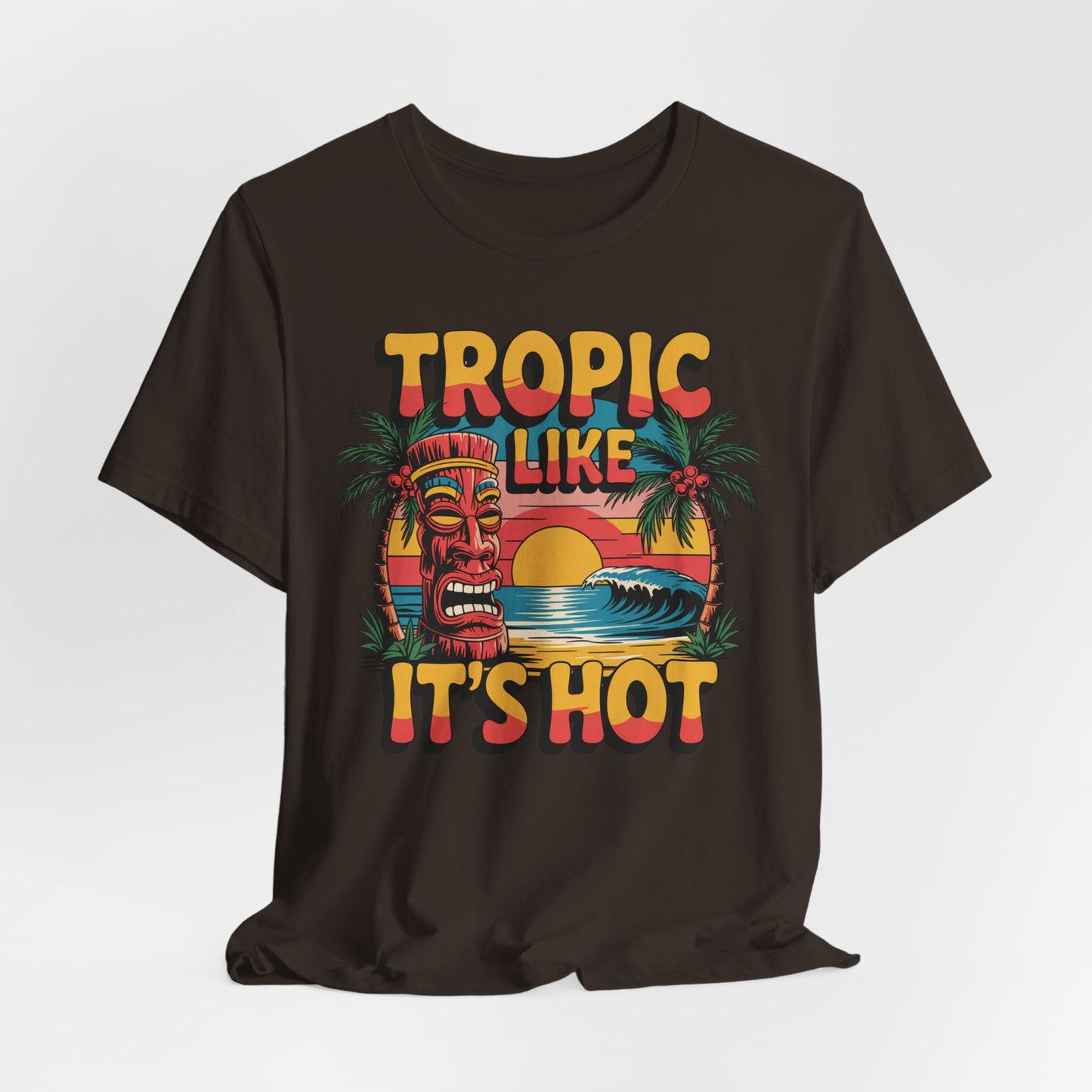 The Tropic Like Its Hot Unisex Tiki T-Shirt features a vibrant tiki mask, palm trees, waves, and a sunset with bold retro lettering—perfect for fun vacation vibes or as your favorite tee for beach days.