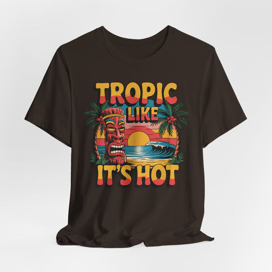 The Tropic Like Its Hot Unisex Tiki T-Shirt features a vibrant tiki mask, palm trees, waves, and a sunset with bold retro lettering—perfect for fun vacation vibes or as your favorite tee for beach days.