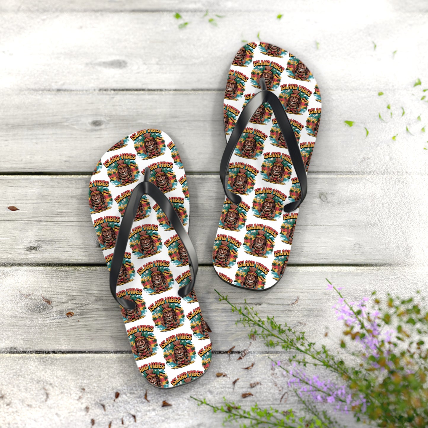 The Island Vibes Tiki Flip Flops feature black straps, vibrant tiki mask soles with “Good Vibes,” and rest on wood beside greenery and purple flowers—ideal colorful casual footwear for summer fun and beach lovers.