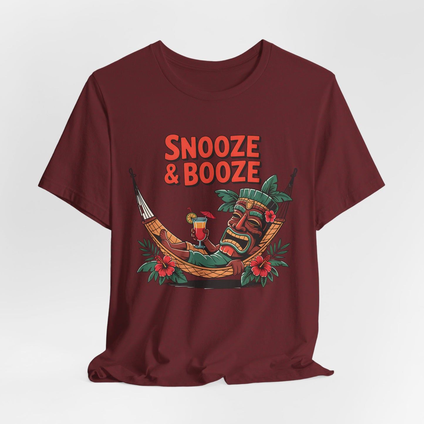 Bring vacation vibes to your look with the Snooze & Booze Unisex Jersey Tiki T-Shirt, featuring Snooze & Booze above a laid-back tiki in a hammock with a drink, lush leaves, and red flowers.