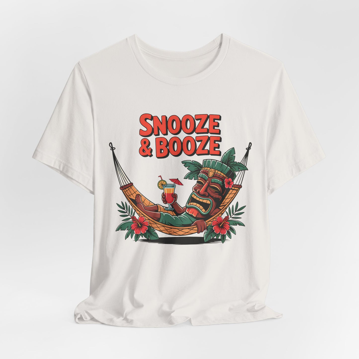 The Snooze & Booze Unisex Jersey Tiki T-Shirt - Tropical Vacation Vibes features a colorful tiki mask in a hammock with a cocktail, bold red SNOOZE & BOOZE text, and tropical leaves and flowers for laid-back, casual style.