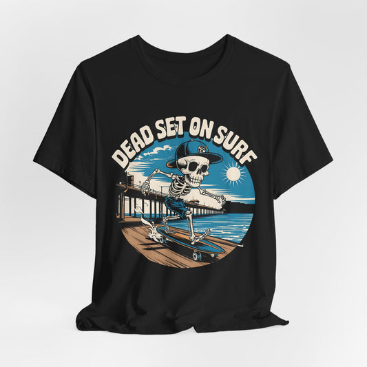 The Skateboard Skeleton Unisex T-Shirt – Dead Set on Surf Design features a bold graphic of a cap-wearing skeleton skateboarding on a pier by the ocean under the sun, with DEAD SET ON SURF above the image.