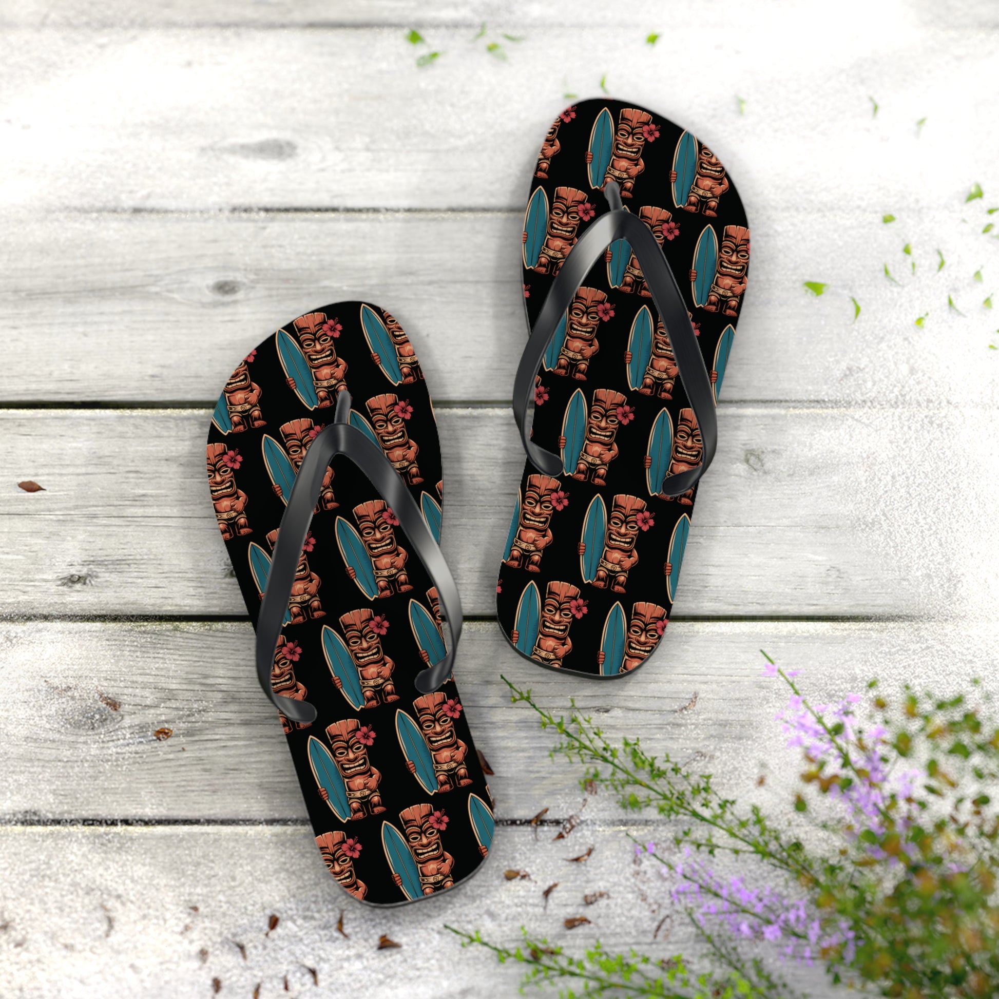 The Tiki Surf Flip Flops - Fun Summer Beach Sandals, featuring black straps and colorful tiki faces with teal surfboards, rest on whitewashed wooden boards amid scattered leaves and purple flowers in the corner.