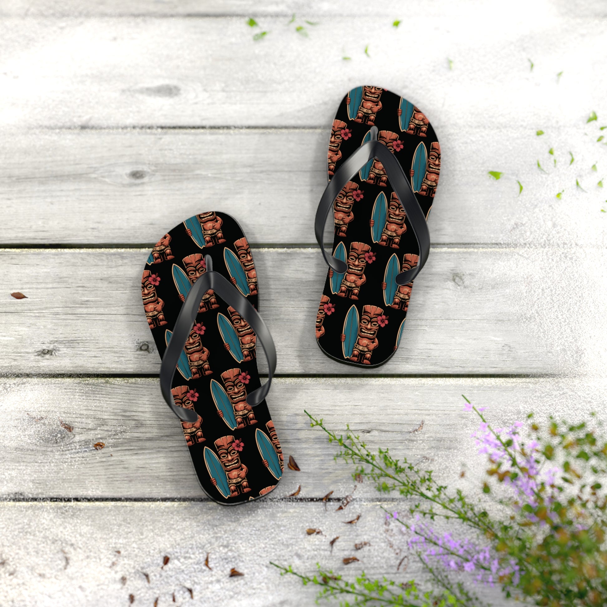 The Tiki Surf Flip Flops - Fun Summer Beach Sandals feature vibrant tiki and surfboard prints on black, shown resting on a light wooden deck scattered with leaves and purple flowers in the corner.