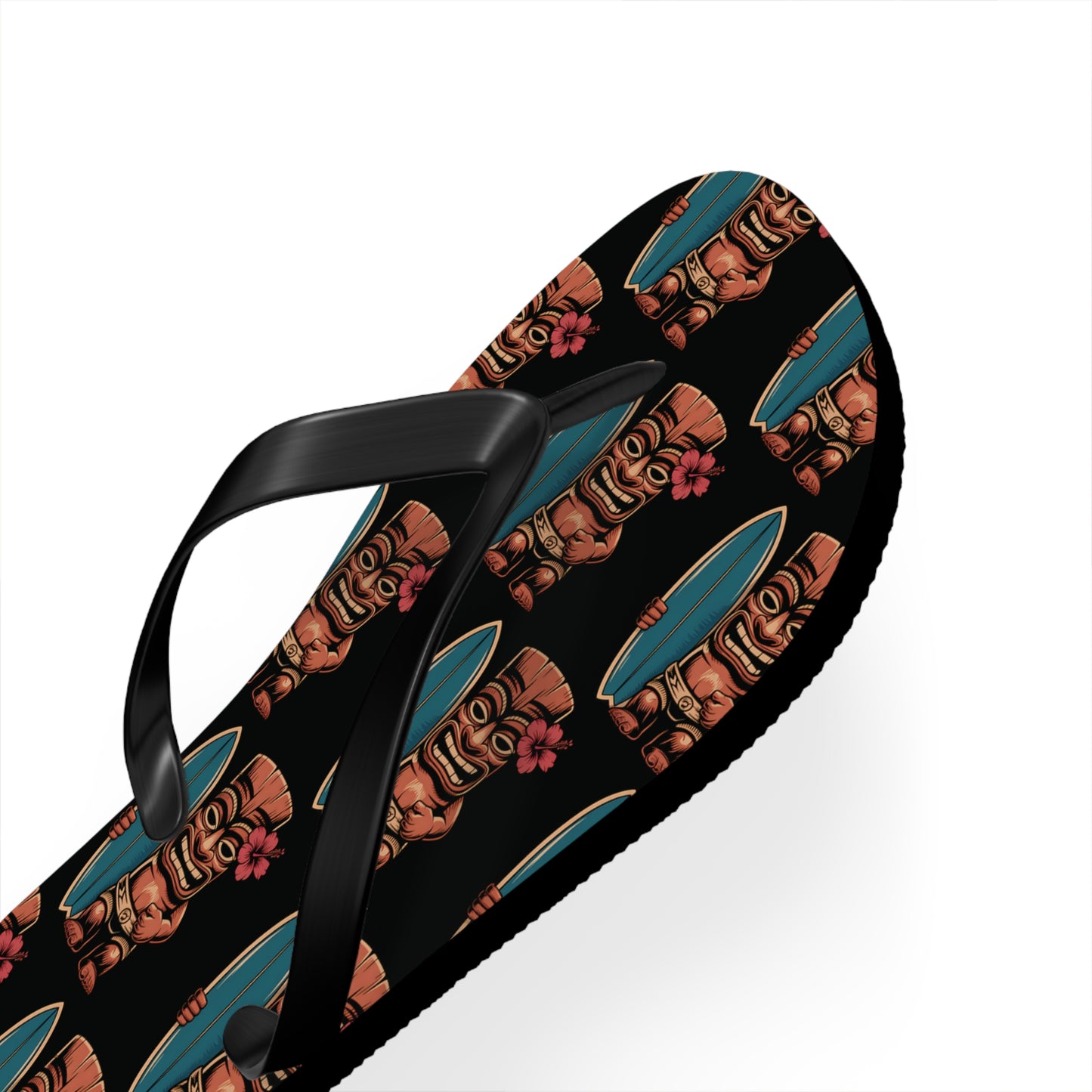 The Tiki Surf Flip Flops - Fun Summer Beach Sandals are black polyester suede flip-flops with vibrant brown tiki faces in blue sunglasses and pink flowers on a white background.