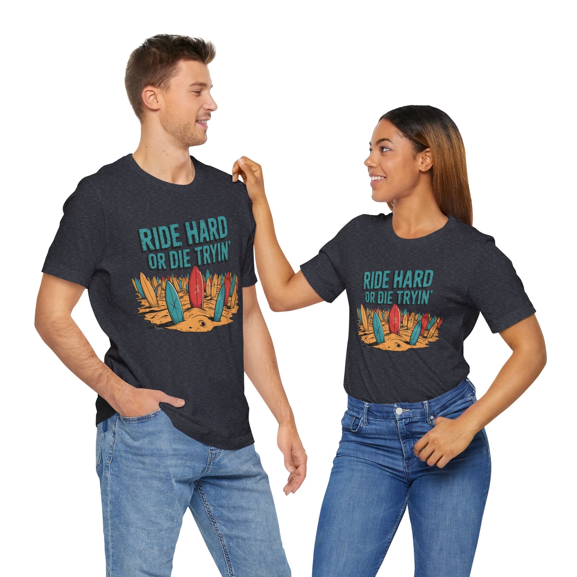 A smiling couple of beach lovers wear matching Surfing T-Shirts – Ride Hard or Die Tryin Unisex Jersey Tees with colorful surfboards and sand, perfect for anyone living the adventure-driven surf lifestyle.