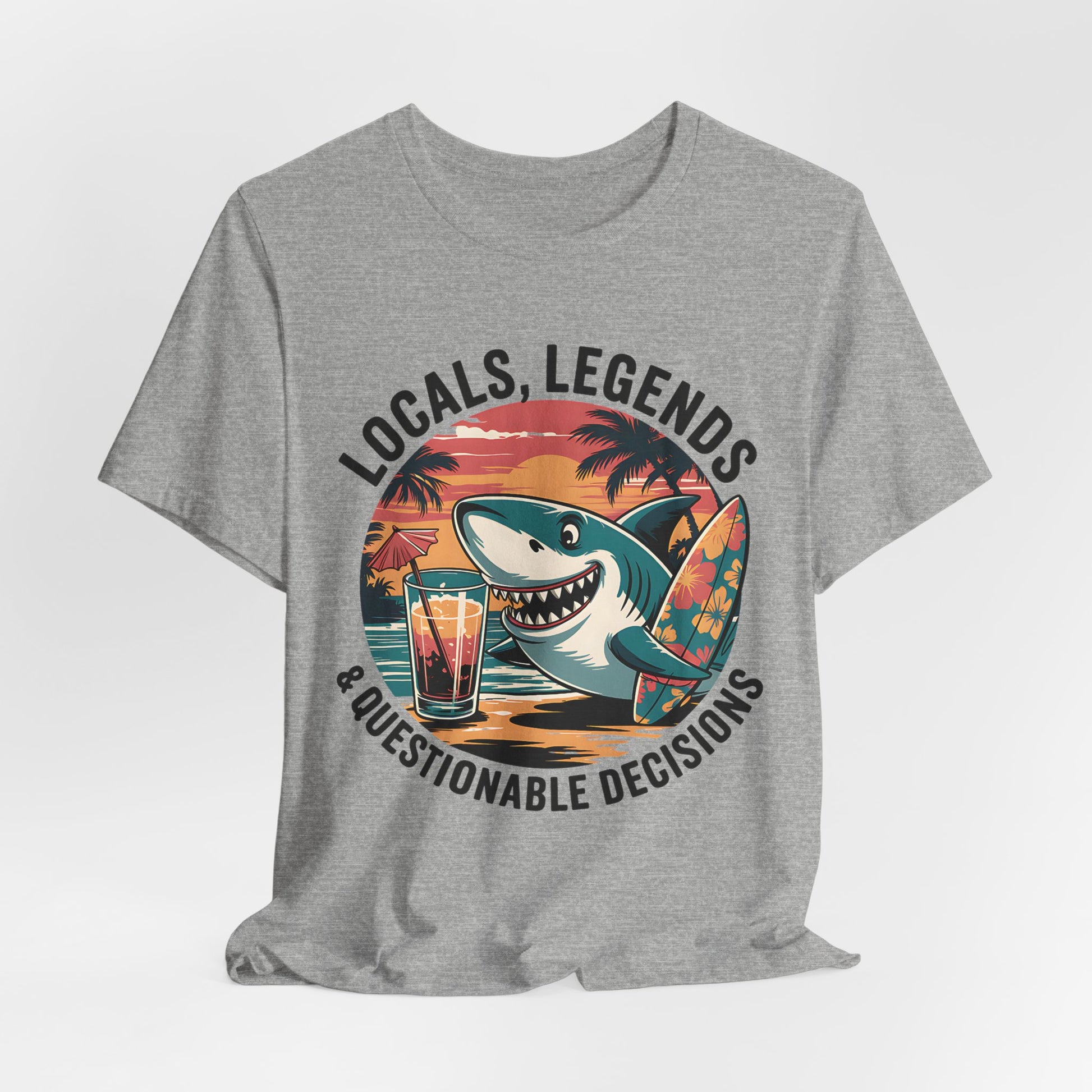 The Funny Shark Graphic T-Shirt - Locals, Legends & Questionable Decisions is a gray unisex tee with a cartoon shark, floral surfboard, cocktail on a table, palm trees, and sunset. Perfect for beach lovers! Text above reads: Locals, Legends & Questionable Decisions.