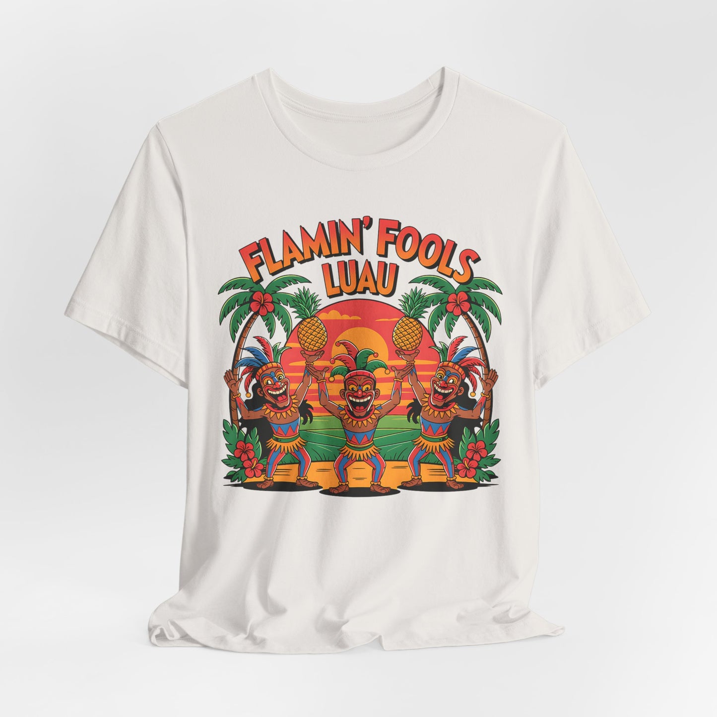 The Flamin Fools Luau Tiki T-Shirt is a white unisex jersey with three cartoon tiki characters holding pineapples, palm trees, and a sunset, plus FLAMIN FOOLS LUAU above. Ideal tropical party shirt for beach gatherings.