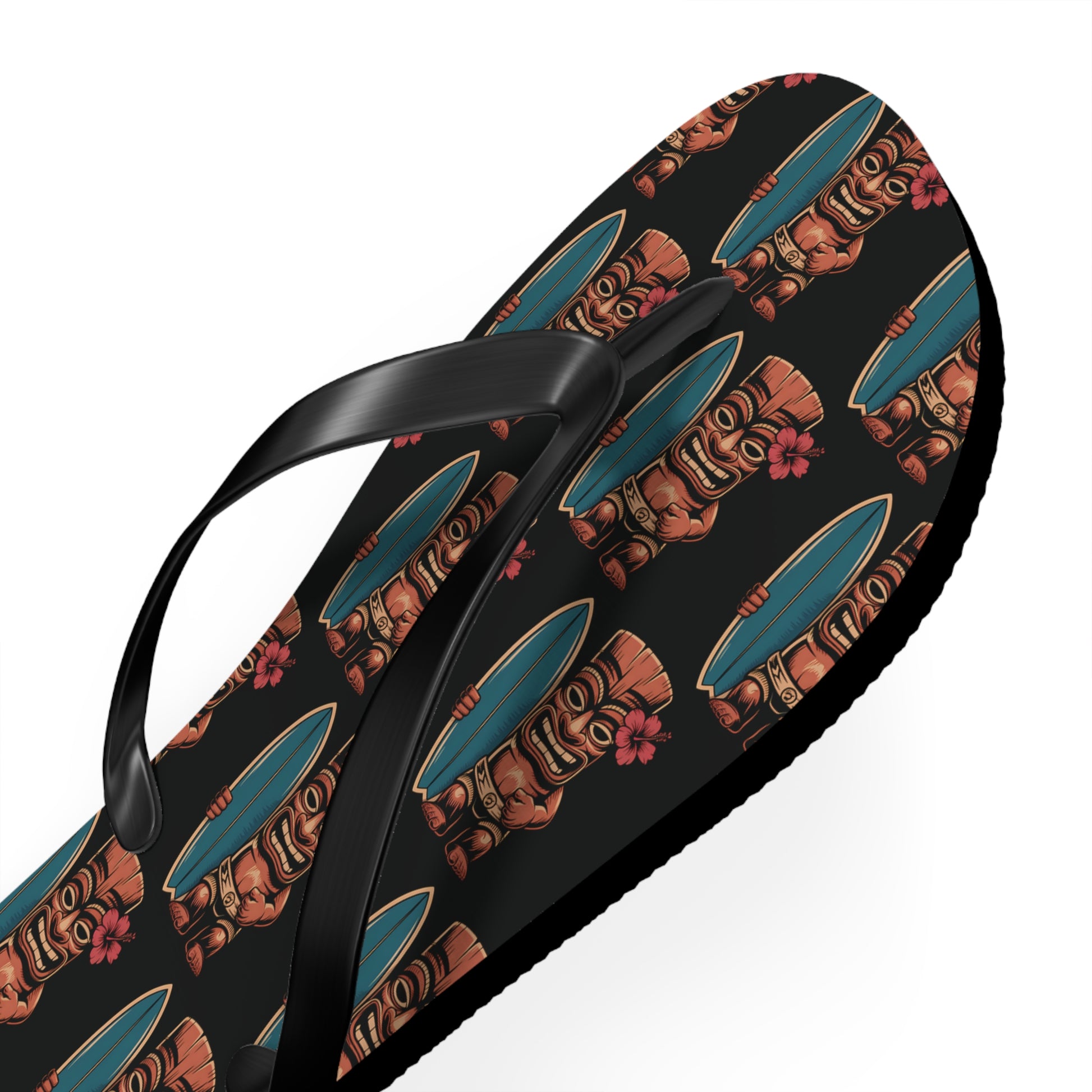 A close-up of the Tiki Surf Flip Flops - Fun Summer Beach Sandals in black, featuring vibrant tiki statues, blue surfboards, and pink hibiscus prints. These flip-flops have black straps, a textured sole, and offer classic summer style.